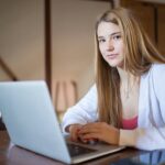 Confident young woman using a laptop while sitting at a wooden desk indoors.