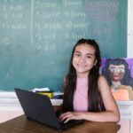 Smiling young student using a laptop in a classroom with a chalkboard background.